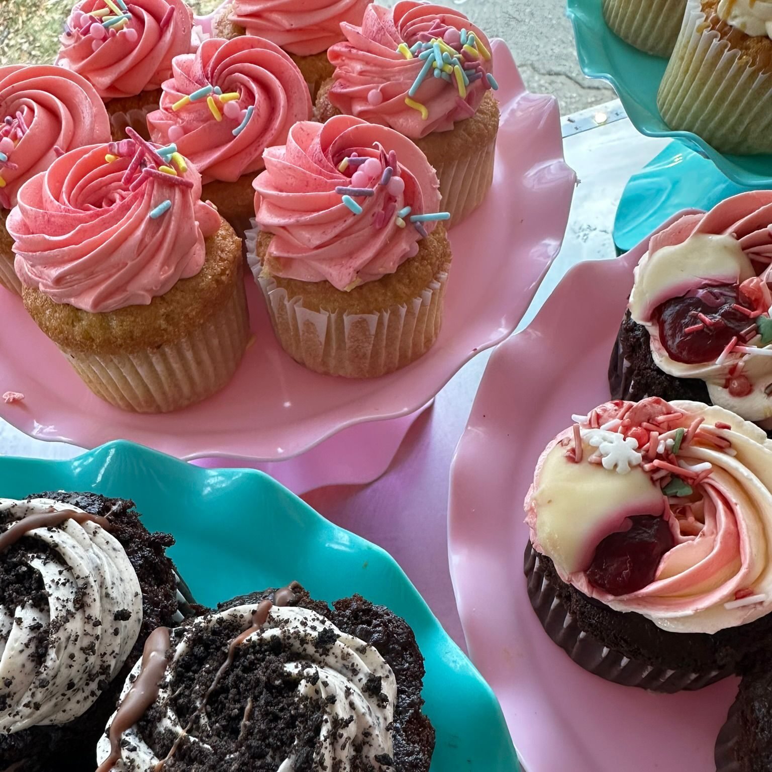 Et utvalg fargerike cupcakes utstilt på pastellfargede fat. Cupcakene er pyntet med rosa og hvit krem, dryss i ulike farger og sjokoladebiter. Noen har mørk sjokoladekakebunn, andre lys vaniljebunn.