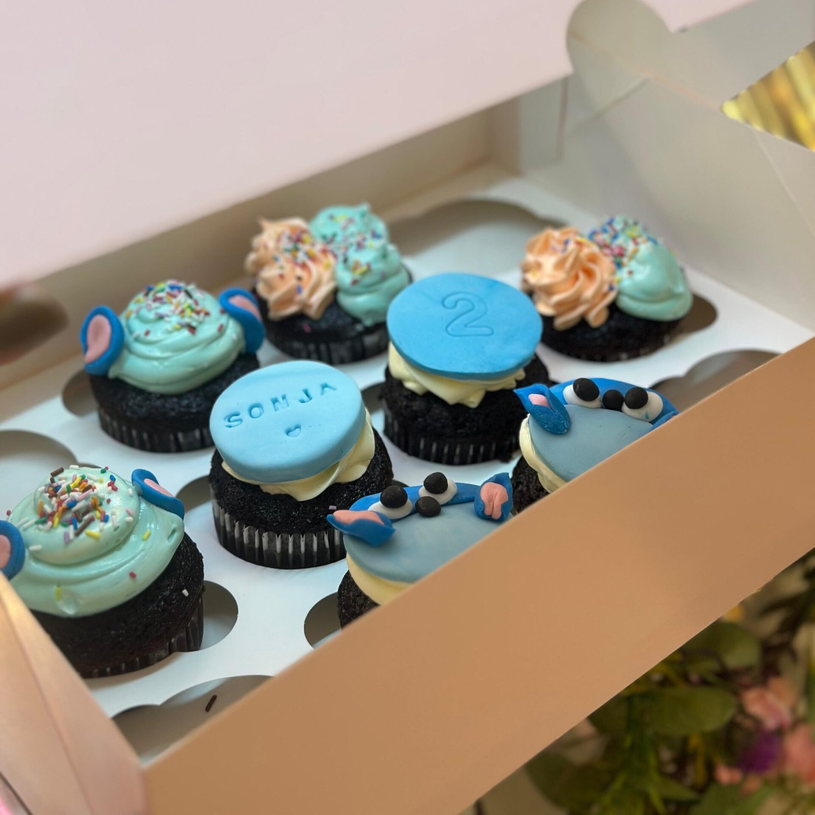 Et utvalg fargerike cupcakes utstilt på pastellfargede fat. Cupcakene er pyntet med rosa og hvit krem, dryss i ulike farger og sjokoladebiter. Noen har mørk sjokoladekakebunn, andre lys vaniljebunn.